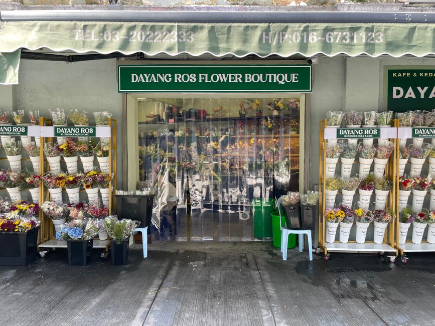 The image shows the exterior of Dayang Ros Flower Boutique on Rush Green Road, with a green sign displaying the shop name. Outside, there are white display shelves filled with various vibrant flower b