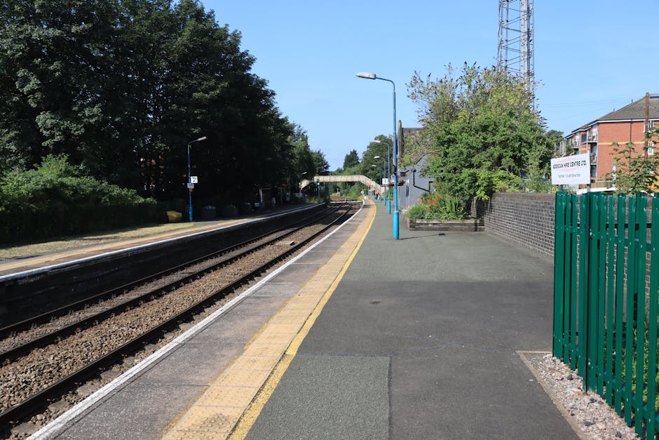 The image depicts the platform at Rush Green station, featuring a concrete surface with a yellow tactile paving strip along the edge for safety. On the right side, there is a green metal fence, behind