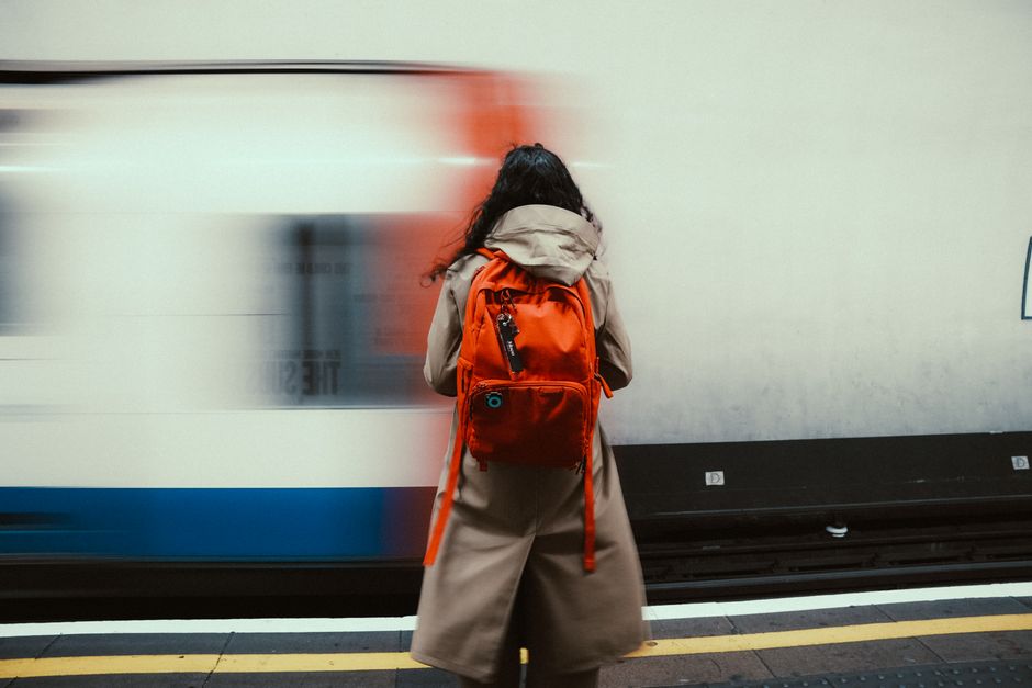 A woman with long, dark, curly hair, dressed in a beige coat, stands on a train platform facing a moving train. She holds a vibrant red backpack on her back. The train is in motion, creating a blurred