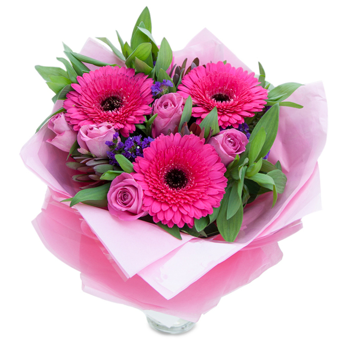 I often think of pink bouquets as holding both mischief and grace; this one shows that balance clearly. Three confident gerbera daisies in a saturated fuchsia dominate the scene, their petals wide and animated around dark, almost velvety centres that read like punctuation marks against the lighter tones. Around them nestle roses in a muted lavender-pink-buds held close in that fresh, unopened stage-each petal layered like a soft whisper. Sprays of purple-blue statice are tucked into crevices between the blooms, offering a rustic, wildflower note and delicate texture that contrasts with the smoother rose petals. Broad leaves-suggestive of salal and sprigs of eucalyptus-fill the background, their green tones cool and dewy, giving the bouquet a sense of rootedness as if recently gathered from a local garden. The flowers are enveloped first in a soft, matte pale pink tissue that creates a gentle base, then wrapped in a glossy yet slightly crinkled translucent fuchsia cellophane which refracts the light and ties the colour story together. The clear glass vase rim shown at the bottom signals the bouquet is ready for easy display. Bathed in soft daylight, the arrangement reads as intimate and celebratory-perfect for a bedside table, a kitchen counter, or as a thoughtful gift after a stroll past Romford Road shops or Rush Green Park. It's the kind of hand-tied piece a Rush Green florist would assemble with care, blending local sensibility with timeless floral craft for birthdays, anniversaries, or just because.